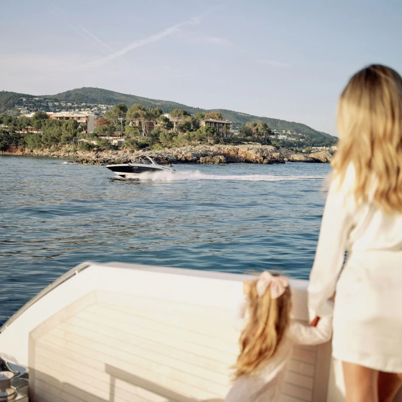 Eine Frau und ein Mädchen stehen auf der Rückseite einer Yacht und schauen auf ein schnell vorbeifahrendes Motorboot auf dem Meer vor einer bewaldeten Küste.