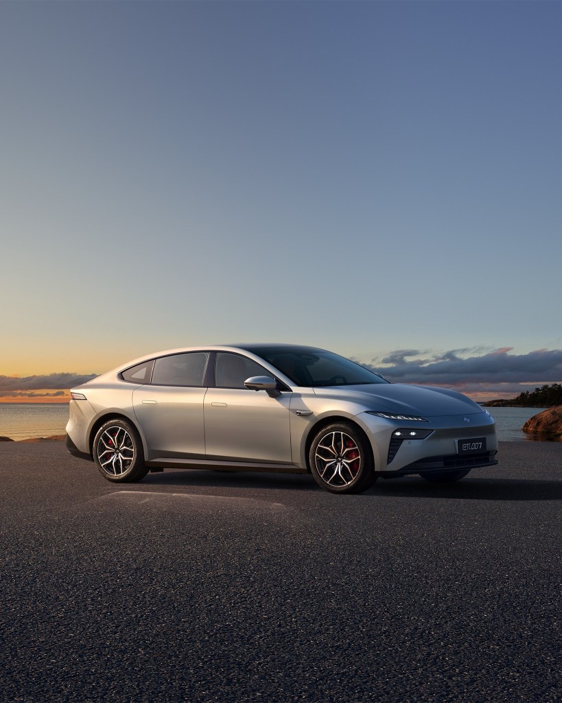 A sleek silver electric car parked on a scenic waterfront during sunset, showcasing its modern design and alloy wheels.