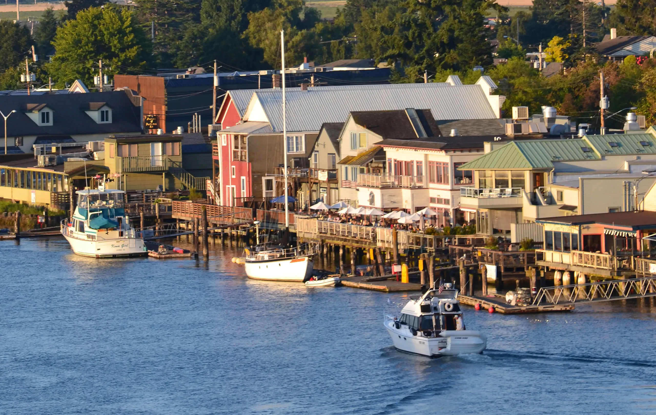 The waterfront of the town of La Conner. There are wooden piers with colorful buildings behind them. Boats are drifting across the water in front of the pier.