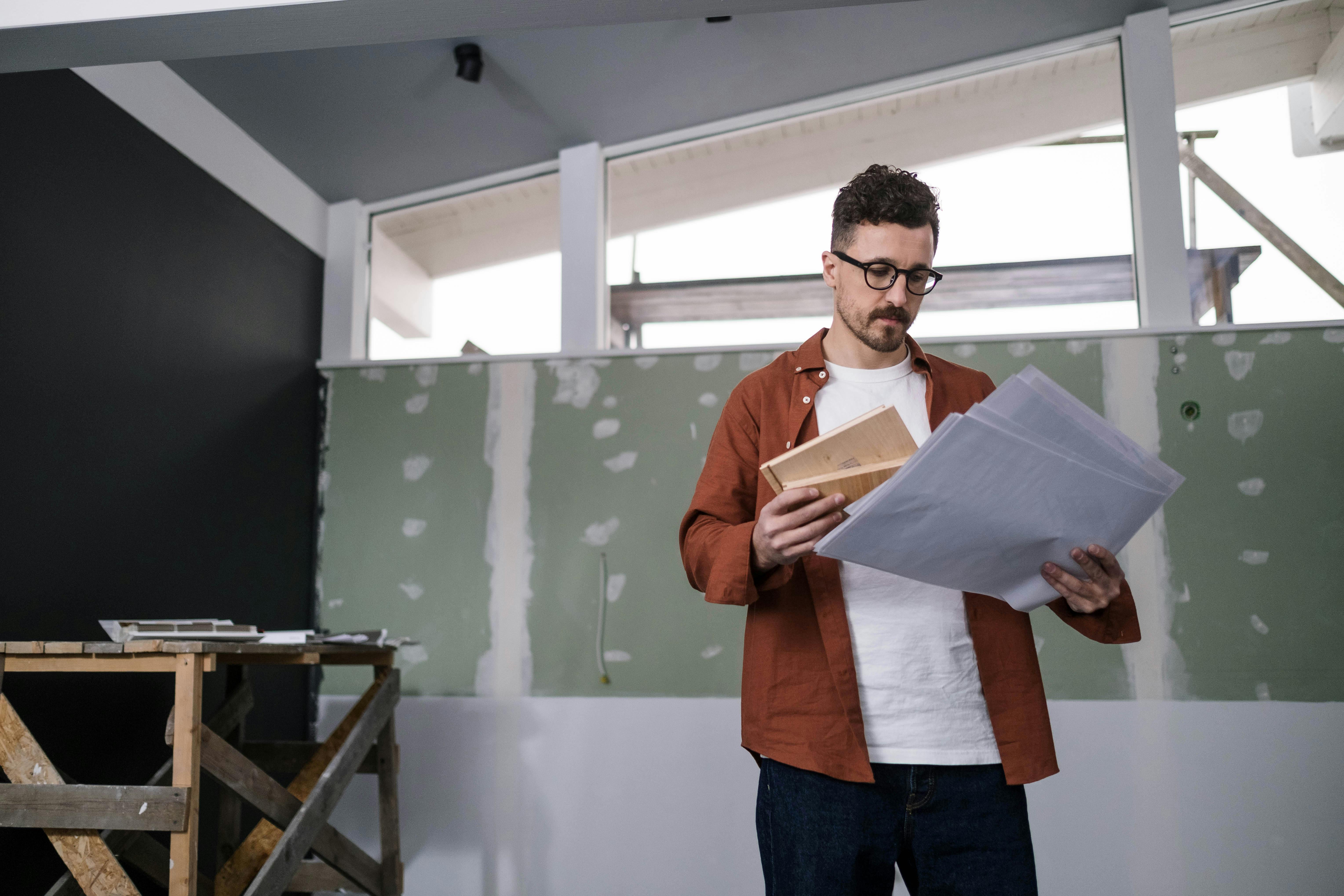 A man in glasses and a brown jacket examines blueprints in a partially renovated room. The setting feels focused and industrious.