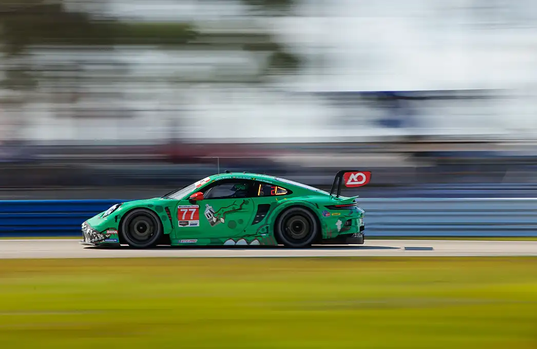 A green Porsche racing car speeds around the Sebring track, with a motion-blurred background.