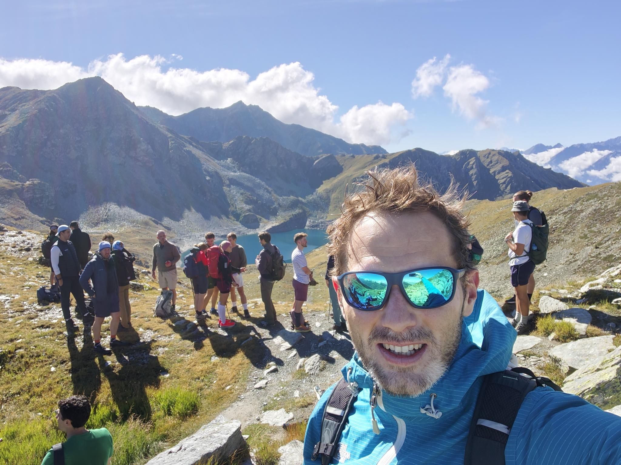 Hiker taking a selfie along a mountain trail overlooking a blue alpine lake.