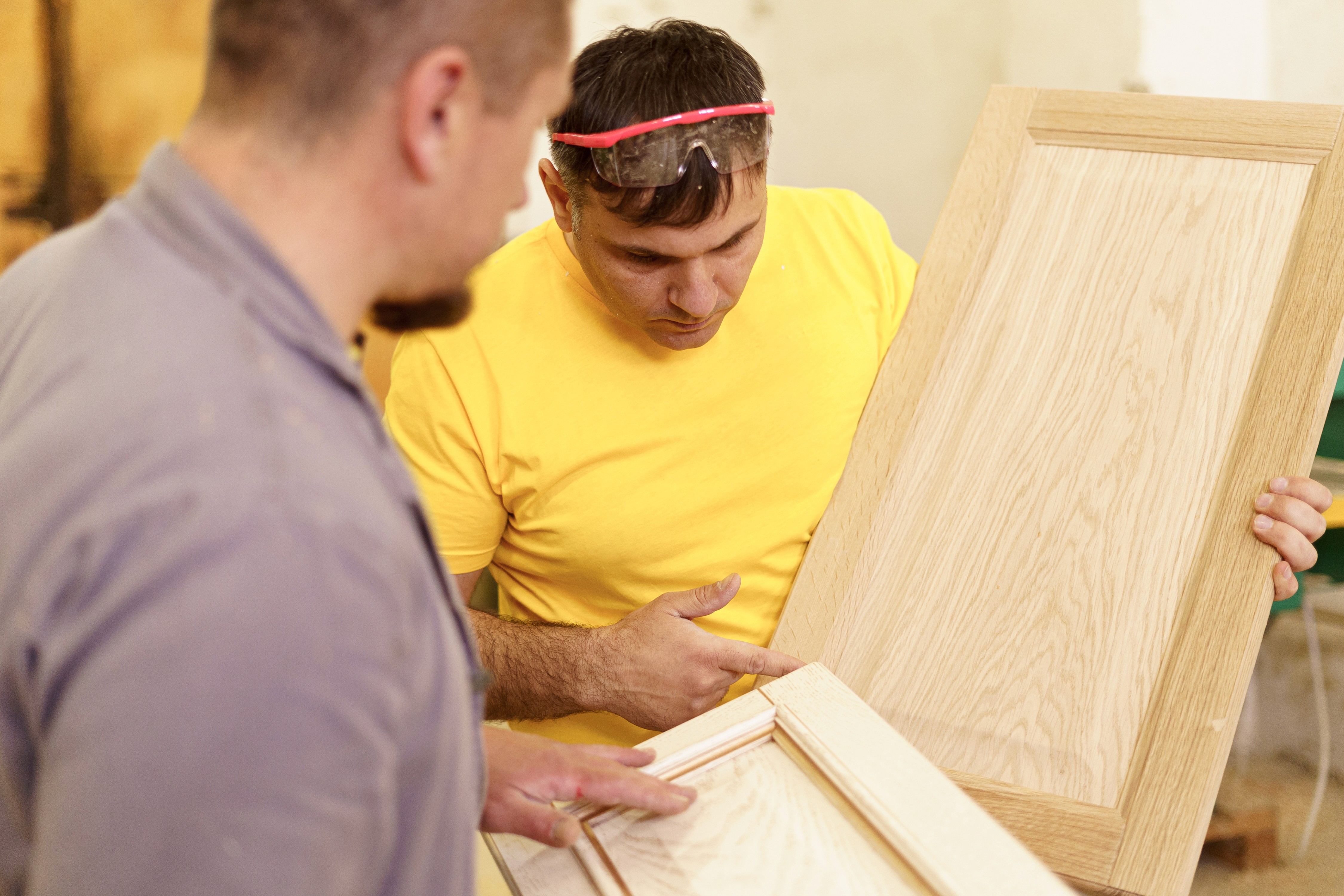 A carpenter wearing safety glasses screws parts of white cabinets together with a drill on a workbench.