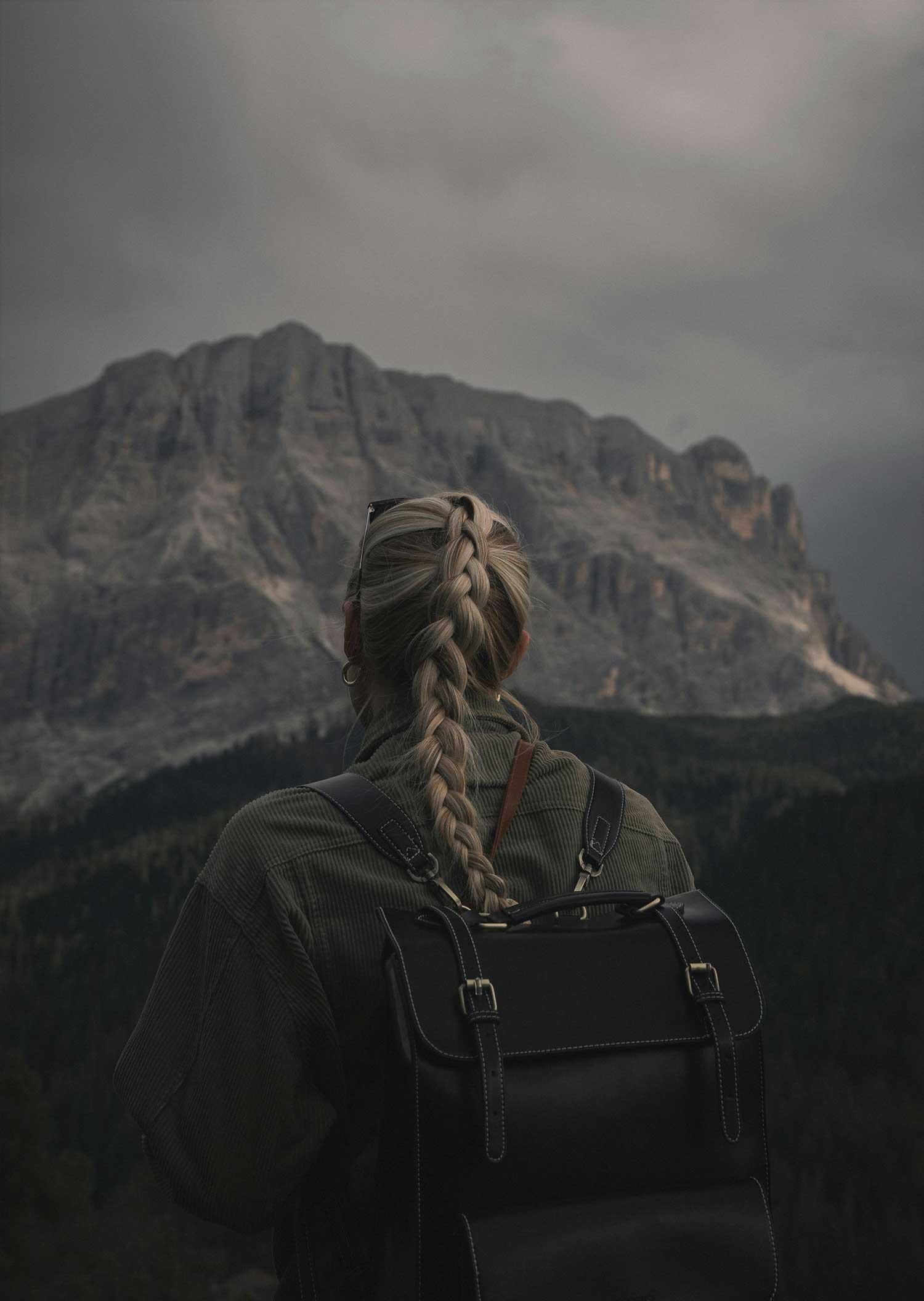 Person with a long braided hairstyle and backpack facing a dramatic mountain landscape under cloudy skies.
