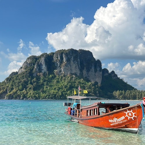 A wooden boat with Thai text is docked in clear turquoise waters, with a large rocky island and cloudy blue sky in the background.