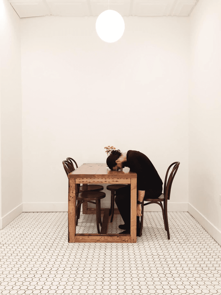 An adult male with long hair dressed in all black sitting at a wooden table in out of four chairs. He's bent at the waist, leaning over the table with his forehead resting on the table. Behind him sits a small vase for several daffodil buds in it.