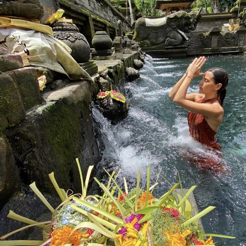 一位穿著紅色禮服的女性在寺廟池塘中祈禱，四周環繞著供品和石雕，水從噴口流出。