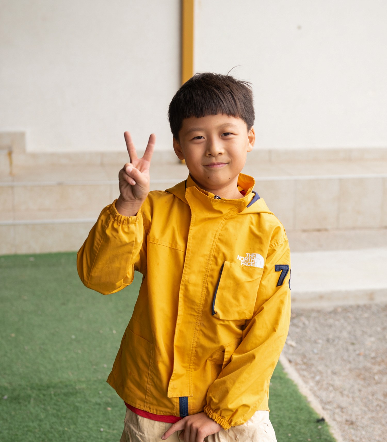 A cheerful child in a yellow jacket smiles and holds up a peace sign indoors.
