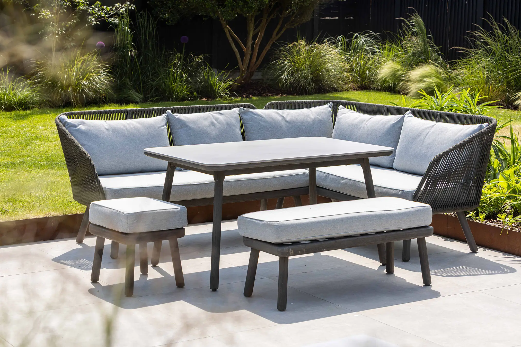 Outdoor seating set with gray cushions, a rectangular table, and a bench, surrounded by greenery.