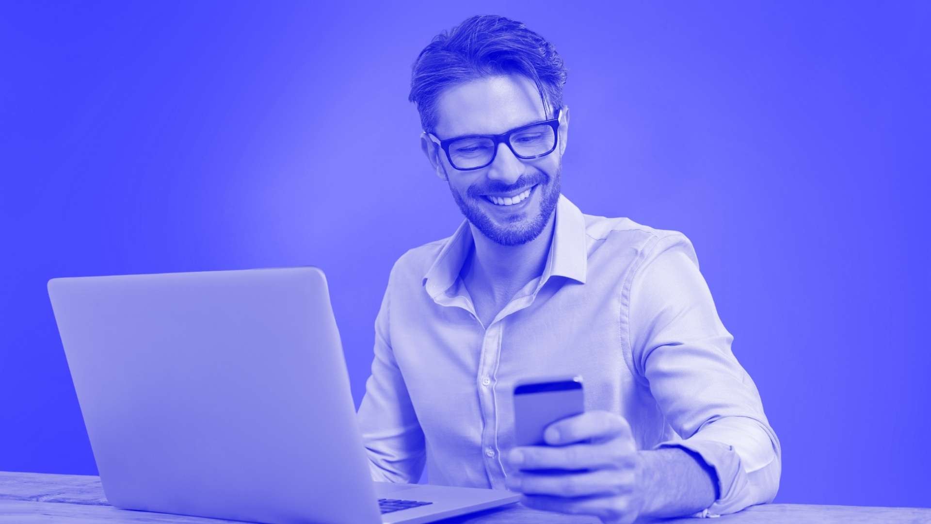 Smiling man wearing glasses sat at a table in front of his laptop looking down at his smartphone