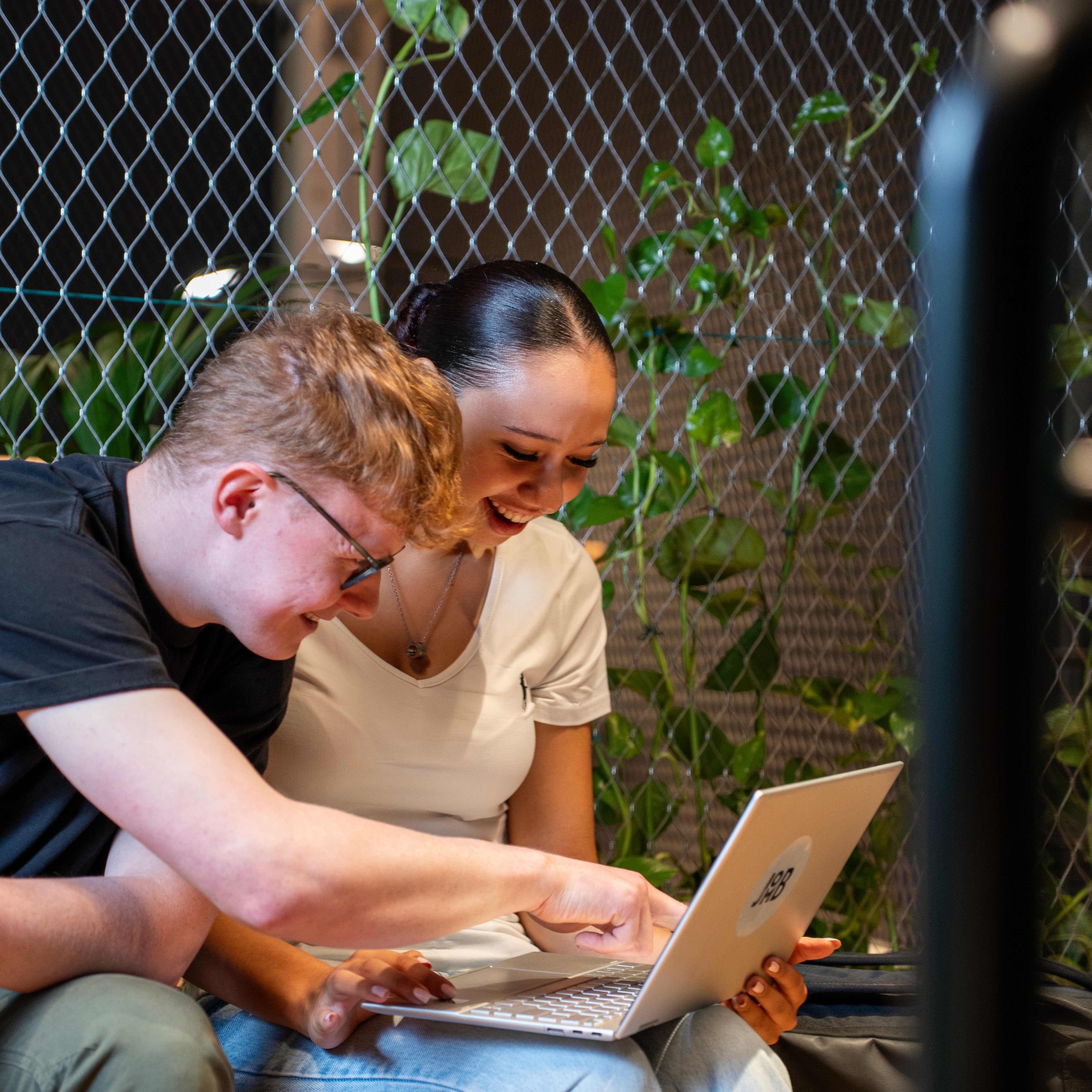 A young person with curly hair smiles at the camera while sitting in front of a laptop against a dark background.