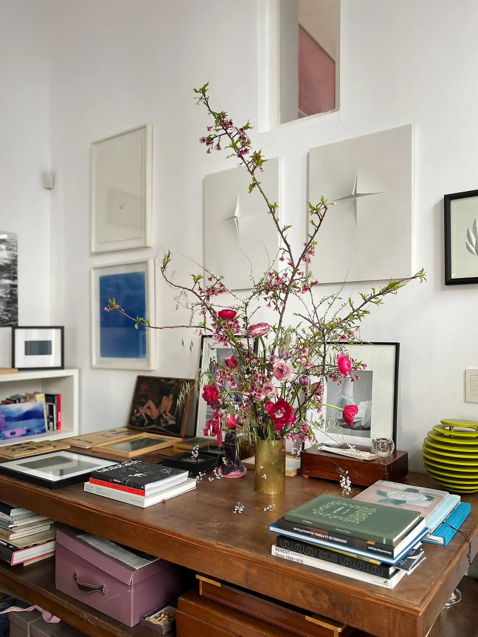 Arreglo floral alto con ramas y flores rosadas en un florero dorado, sobre un escritorio de madera lleno de libros y cuadros enmarcados en un ambiente artístico.