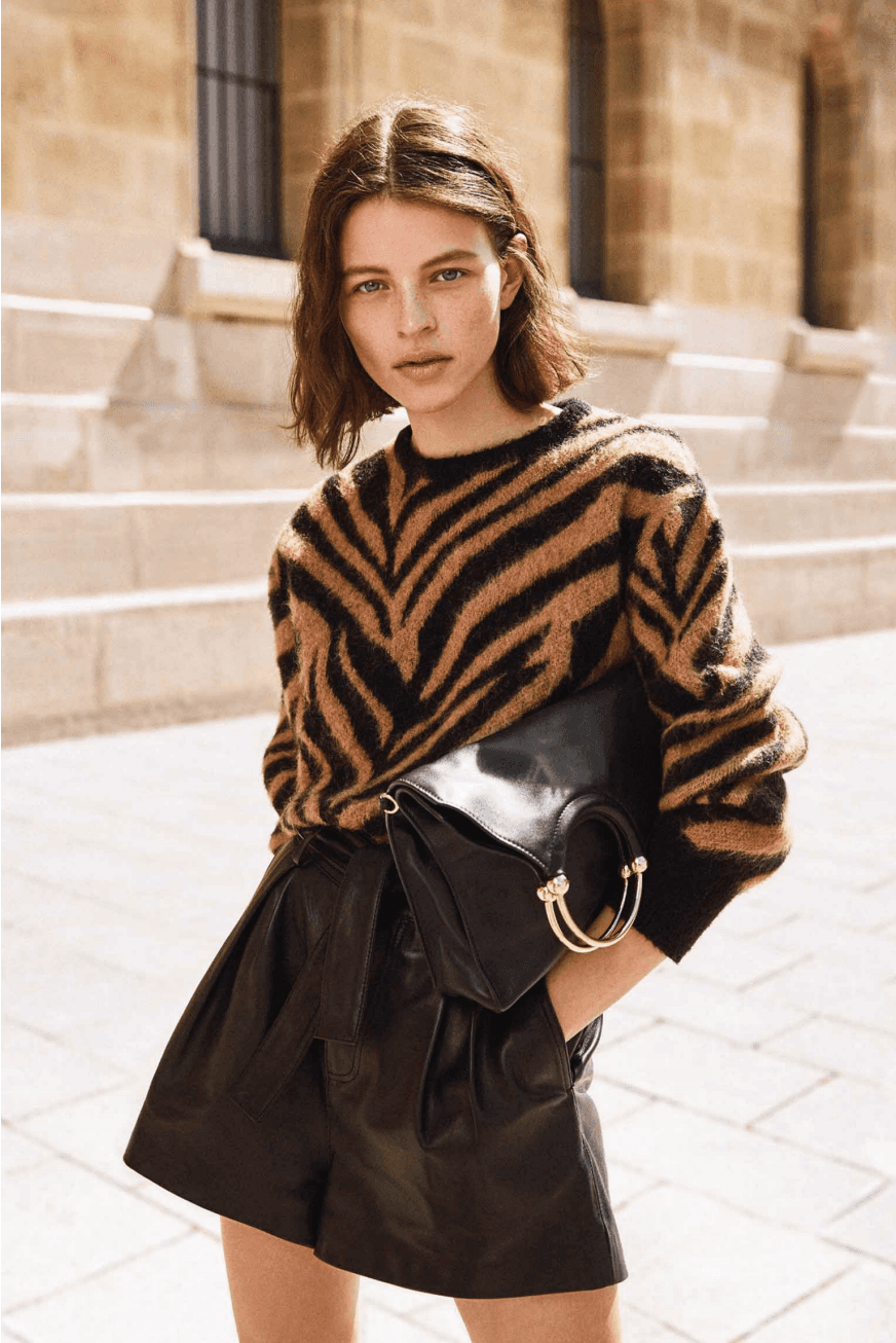 A person with shoulder-length hair is wearing a tiger print sweater and high-waisted black leather shorts, holding a black clutch with a gold accent, standing on sunlit stone steps.