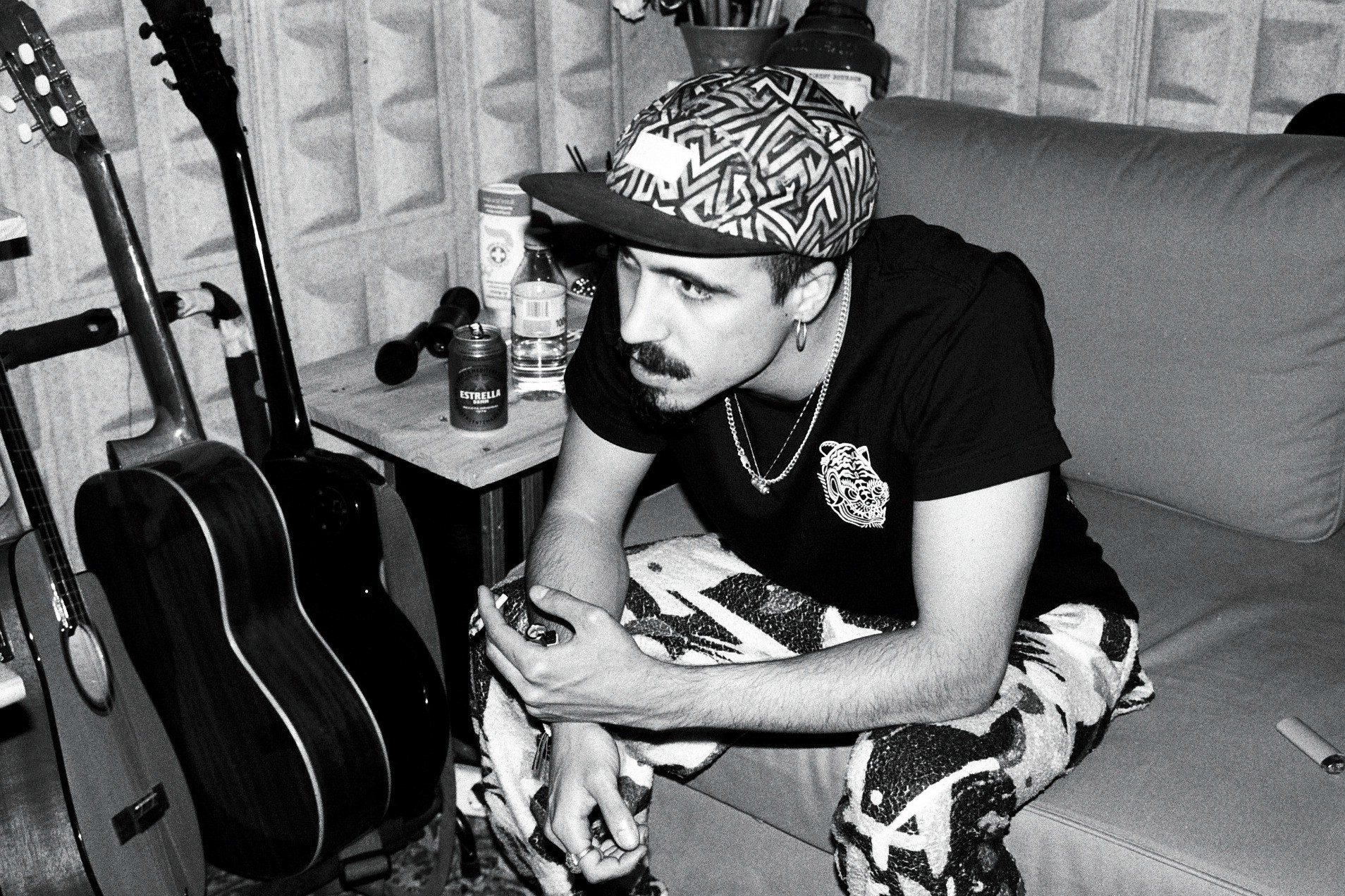 A man in a hat sits pensively in a music studio surrounded by guitars, captured in black and white.