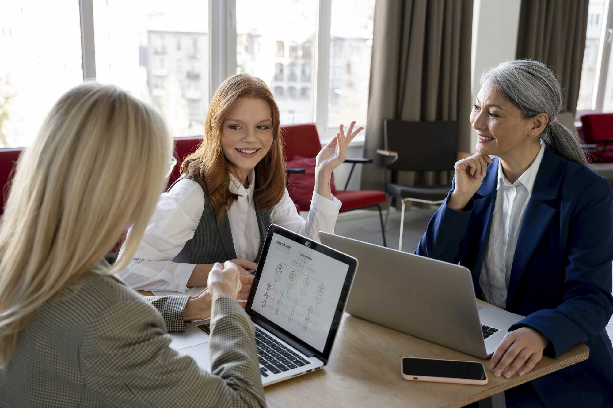 Three professional women collaborating in a bright meeting room, two using laptops and one gesturing during a discussion