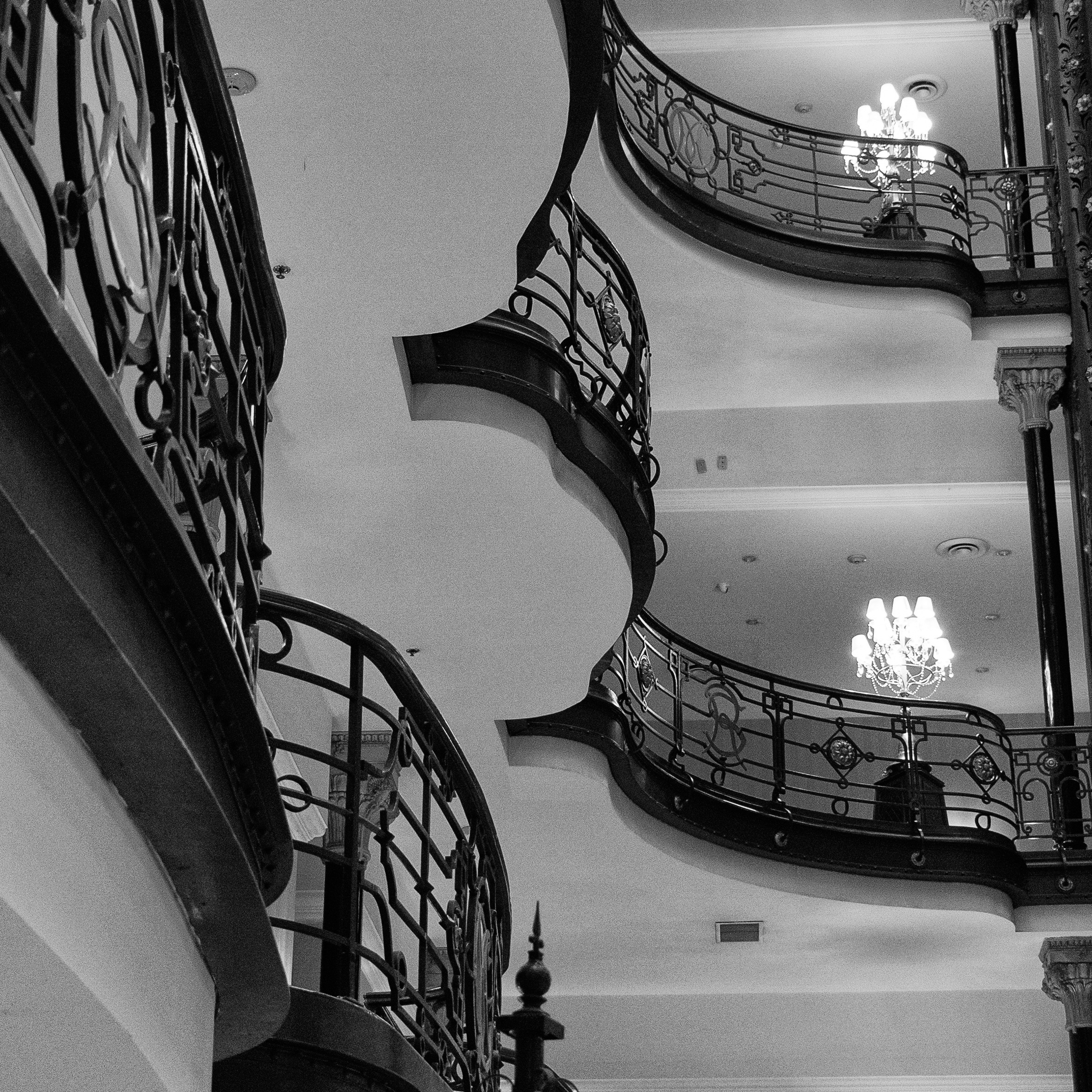 Black and white interior shot of Hotel México showing ornate iron balconies, soft-lit chandeliers, and curved architectural lines.