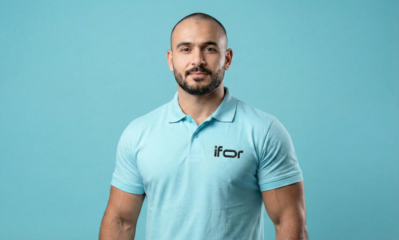 A smiling man with dark hair, wearing a black shirt, poses against a soft blue background.