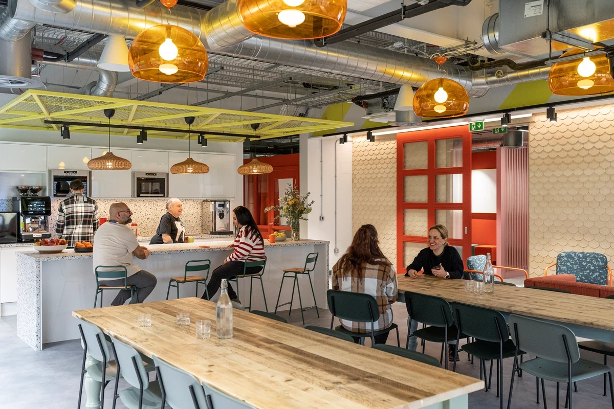 A modern office social space with long wooden tables, a kitchen bar, and pendant lighting, where small groups sit and talk in an open, informal setting.