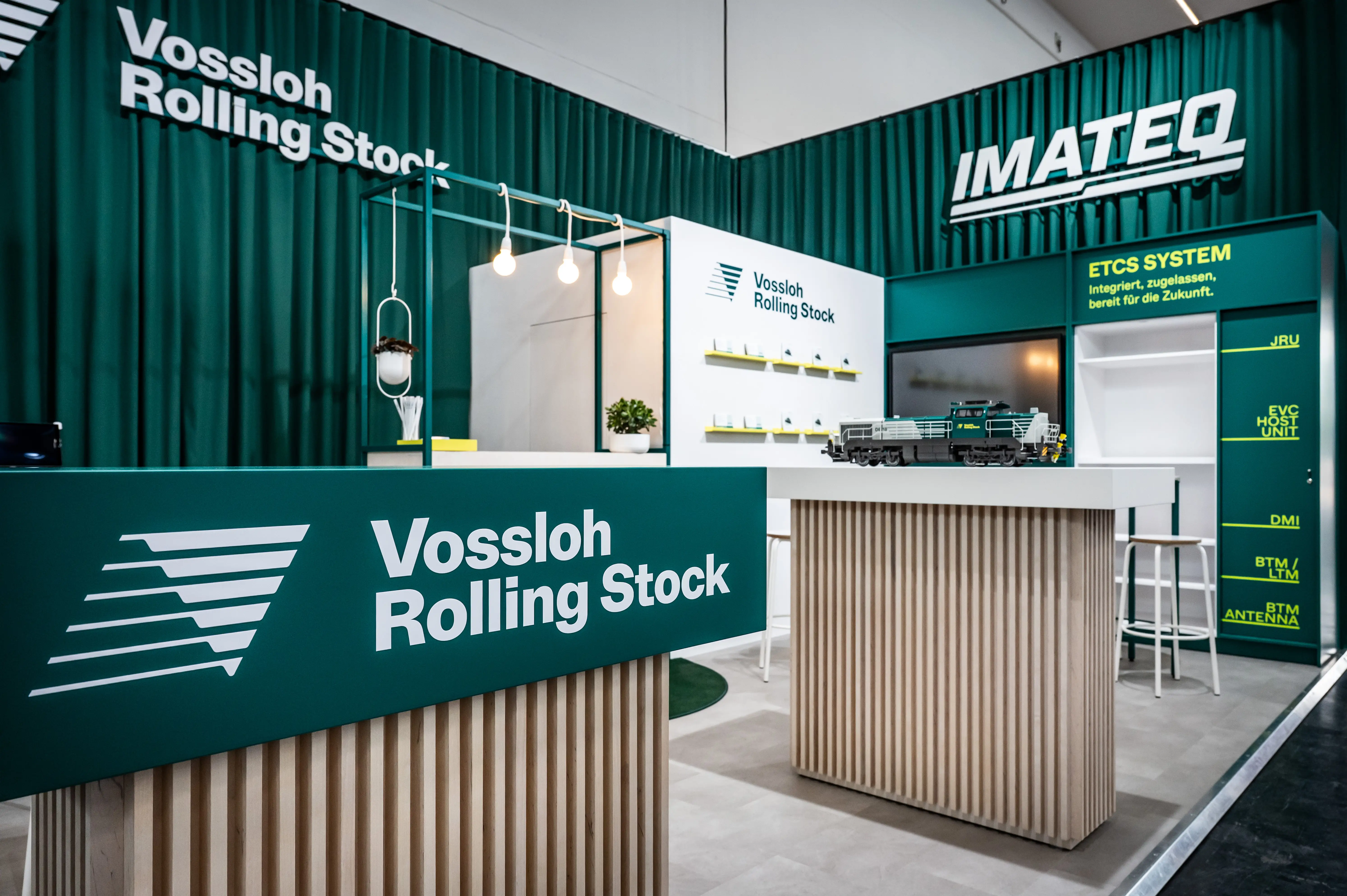 Overview of the Vossloh Rolling Stock booth with green curtains, wooden slat counters and Imateq wall.