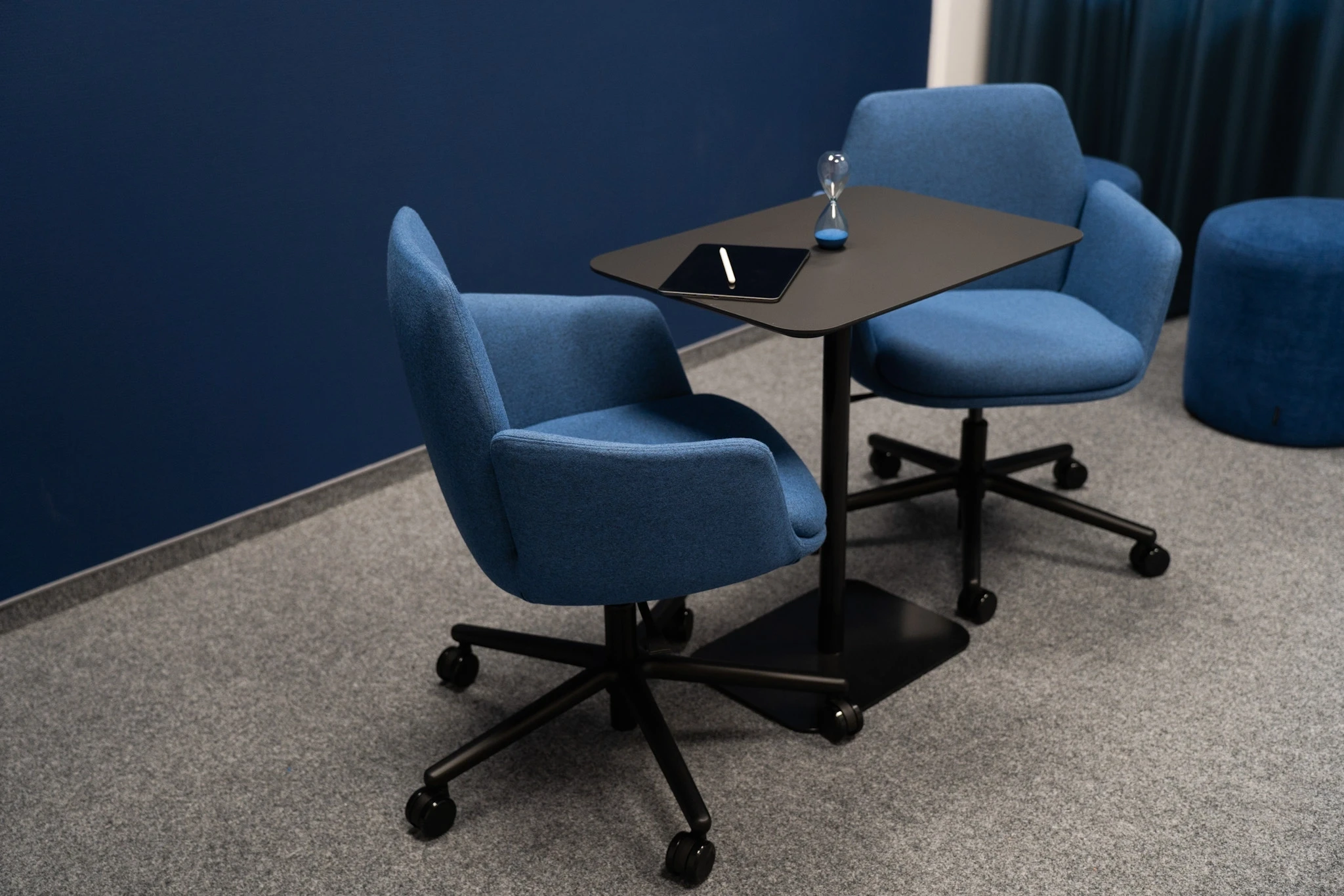 Small meeting corner at NFON with two blue upholstered swivel chairs around a black square table with tablet and hourglass, against a dark navy wall with grey carpet.