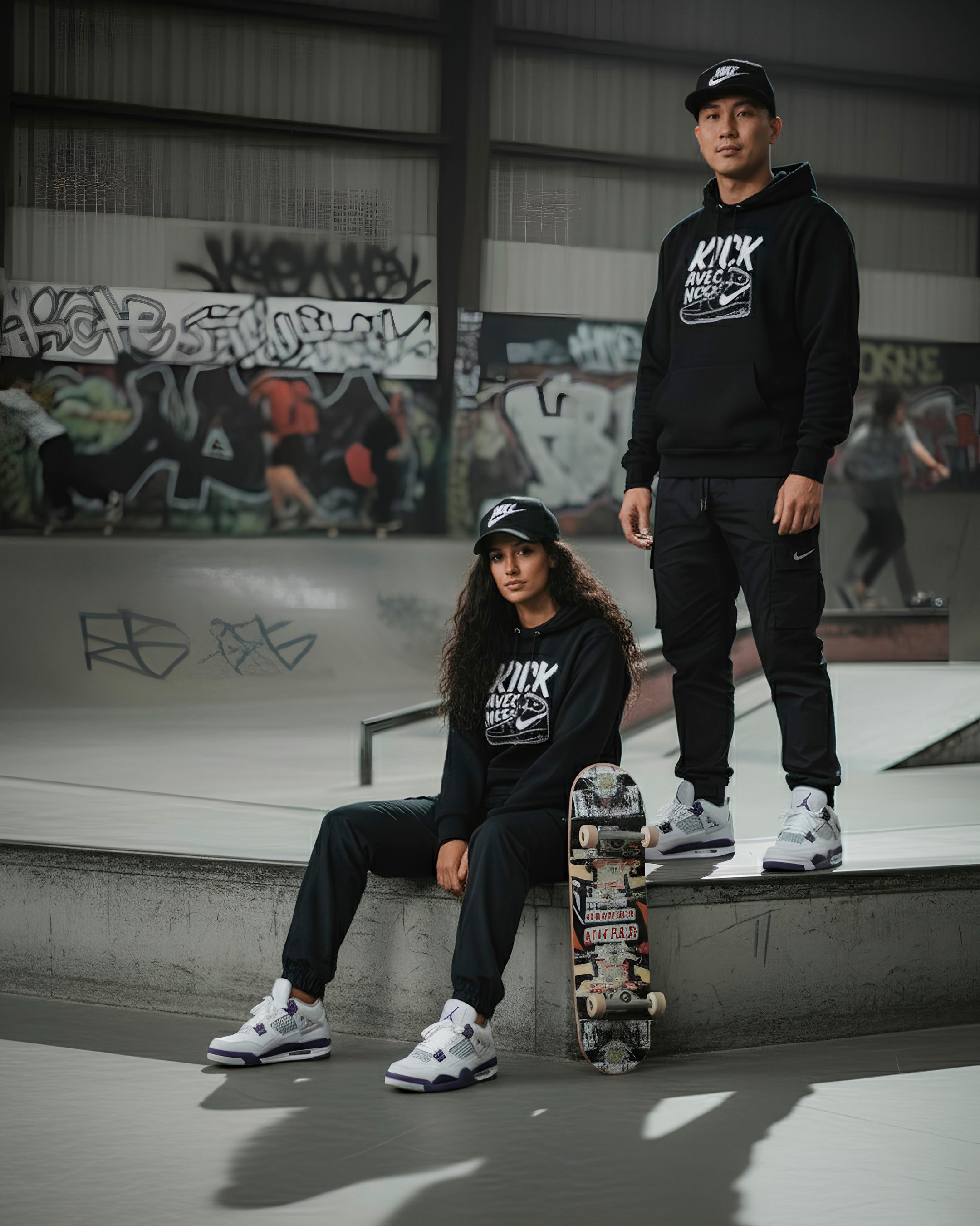 Photo de mode lifestyle d'un duo (homme et femme) posant dans un skatepark couvert et graffité. La femme est assise sur le rebord d'une rampe, tenant une planche de skateboard décorée, tandis que l'homme est debout, les mains à ses côtés. Les deux portent des casquettes noires 'Nike', des sweats à capuche noirs assortis, des pantalons de survêtement/cargo noirs, et des sneakers blanches et violettes (Jordan 4). L'ambiance est urbaine et sombre.