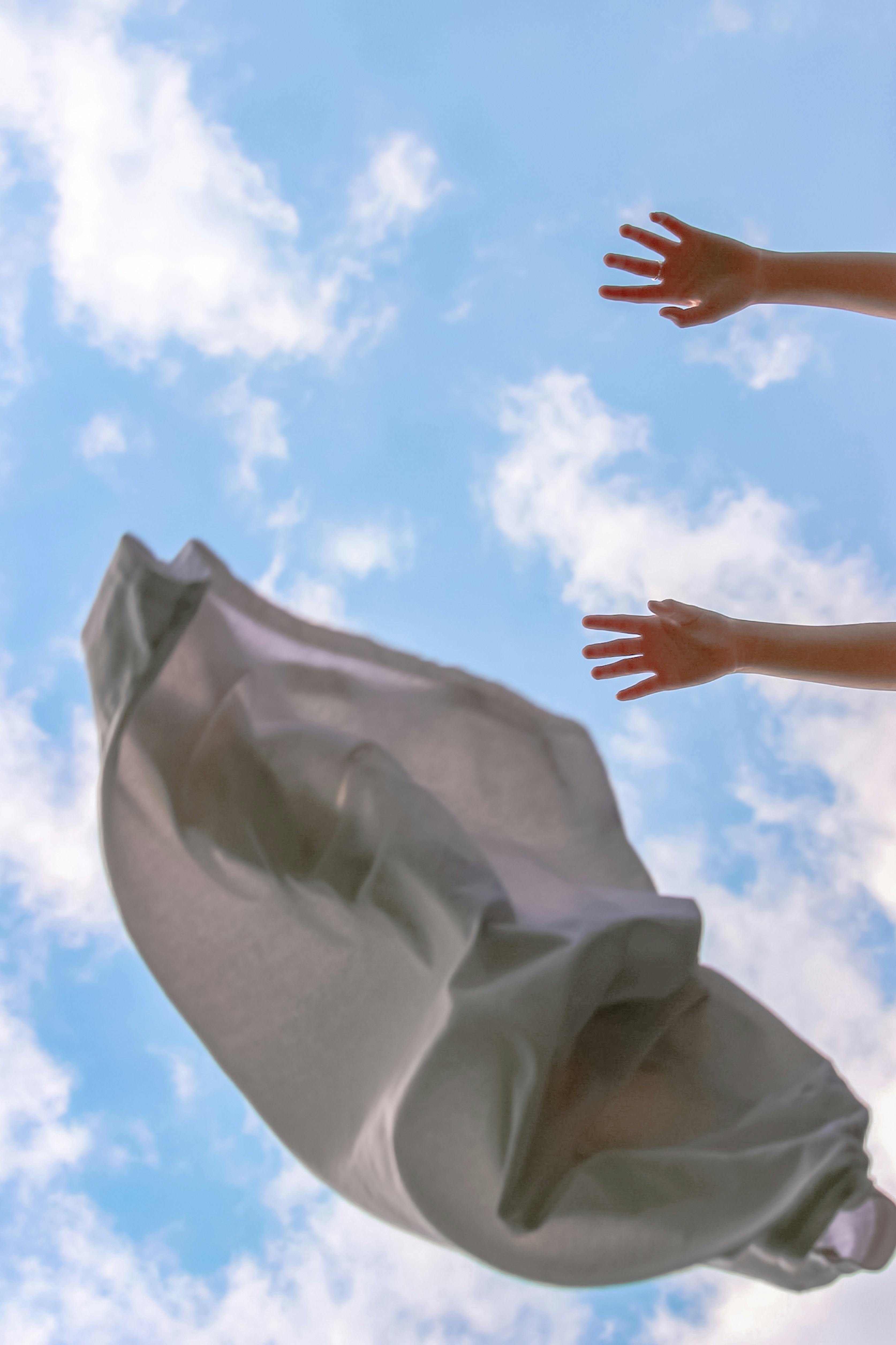 A grey linen garment held up against a bright blue cloudy sky with an outstretched hand