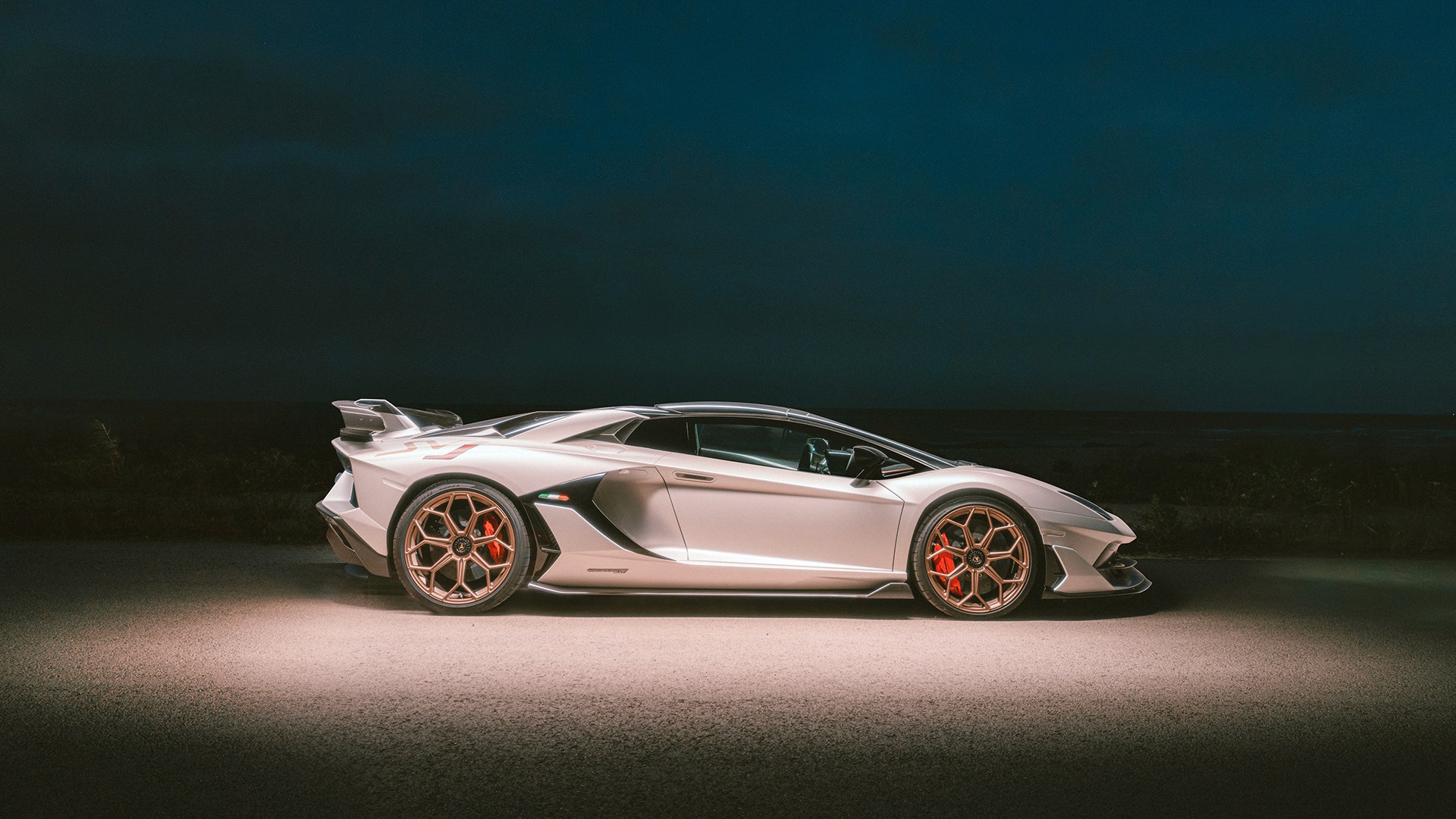 White Lamborghini Aventador SVJ with bronze wheels in side profile, lit against a dark blue night sky, showing the high rear wing, side air intakes, red brake calipers and "Aventador SVJ" sill script, parked on open ground.