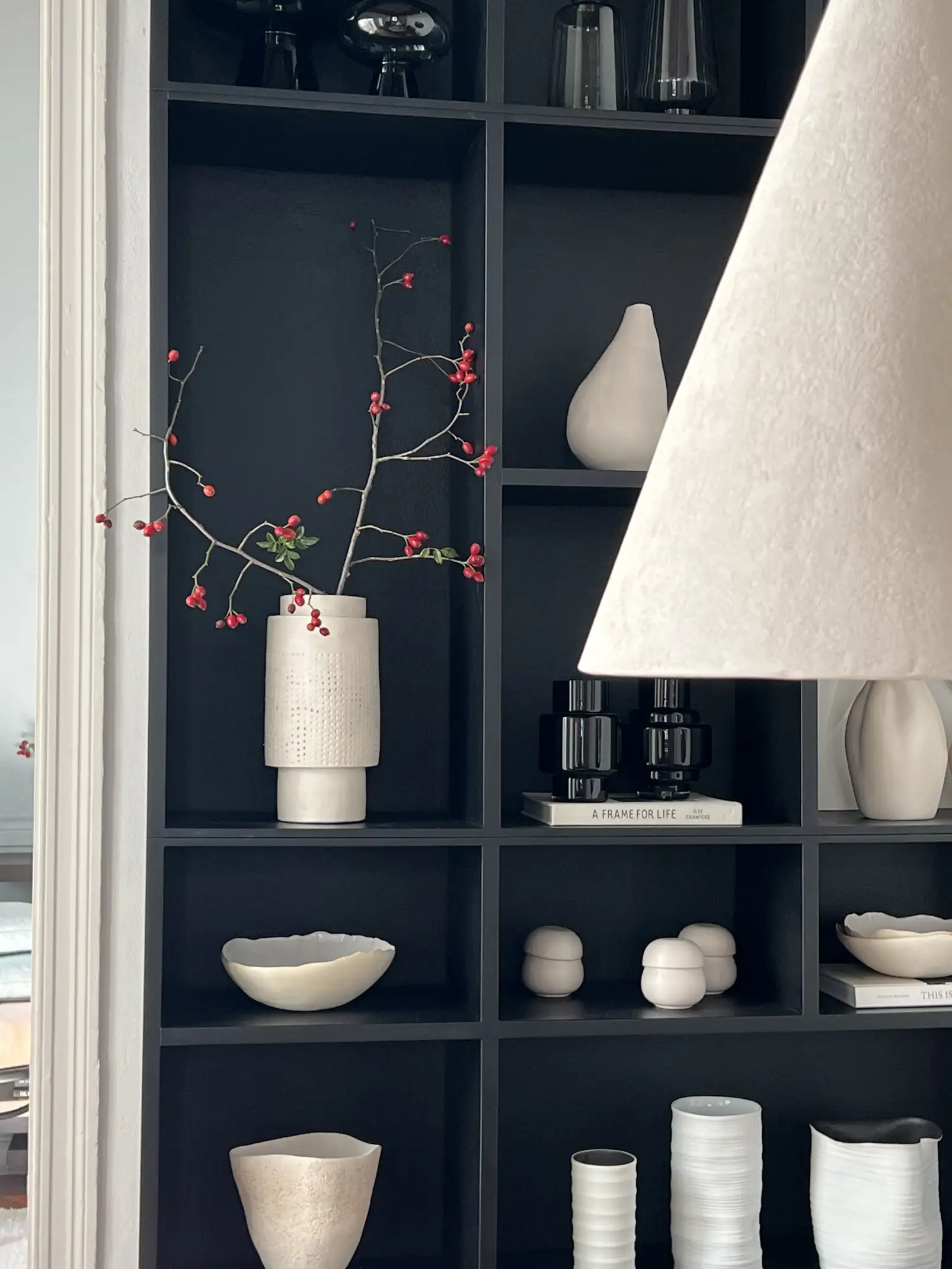 Estantería negra con cerámicas blancas y arreglo con ramas rojas para estilistas de interiores que destacan contraste y composición.