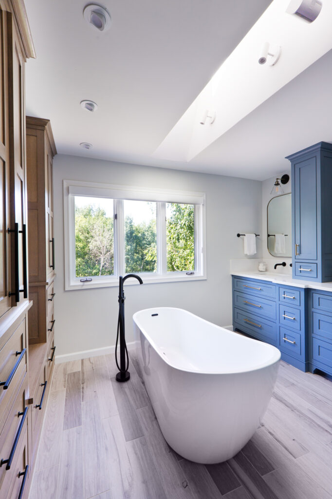 Bright modern bathroom featuring a white freestanding tub, light wood floors, large skylight, and built-in wooden and blue cabinetry. A walk-in glass shower with black fixtures sits near a private toilet room.