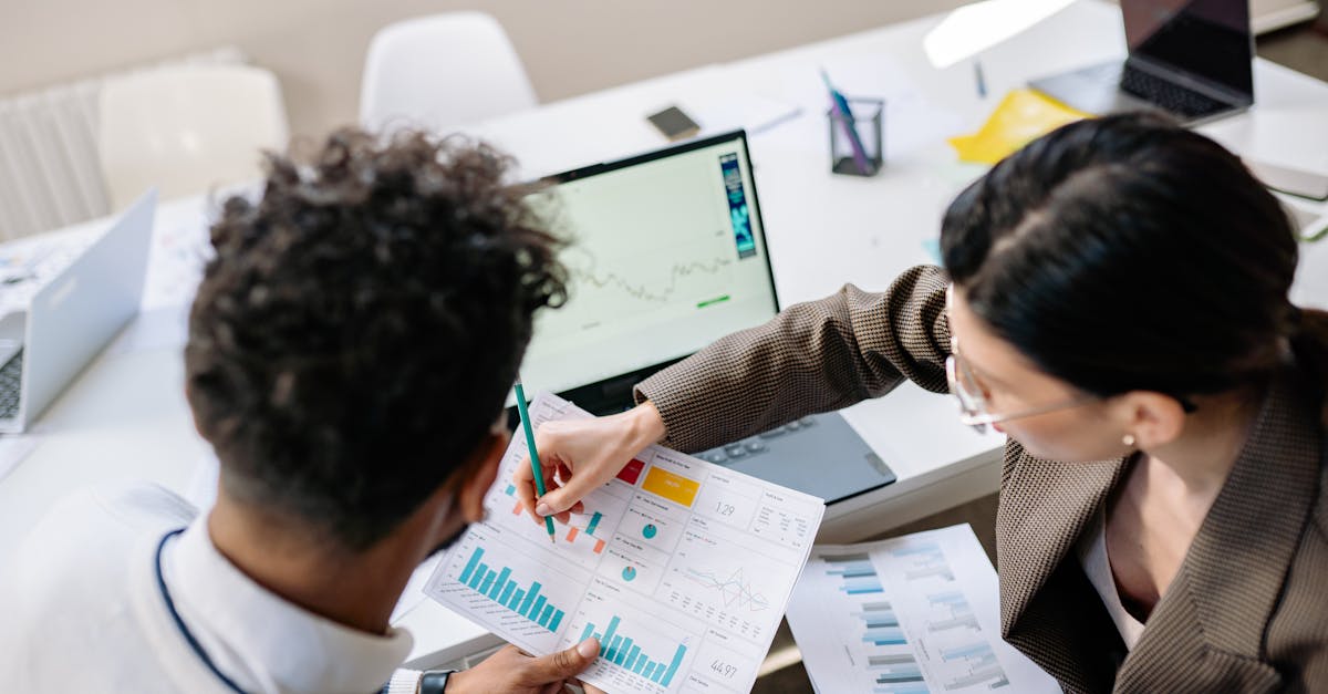 Two colleagues in a meeting room discussing financial charts and graphs on a laptop and paper.