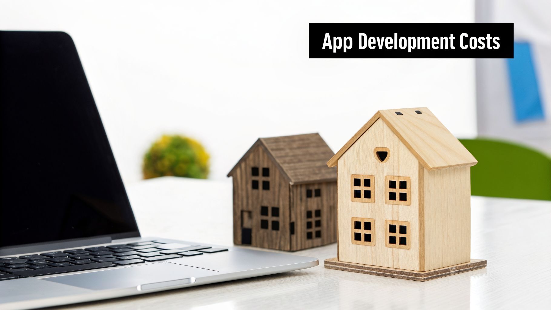 Laptop and two miniature wooden houses on a white desk with 'App Development Costs' banner.