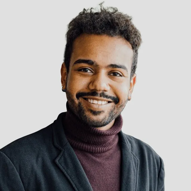 Smiling man with short curly hair wearing a dark blazer and maroon turtleneck sweater poses.