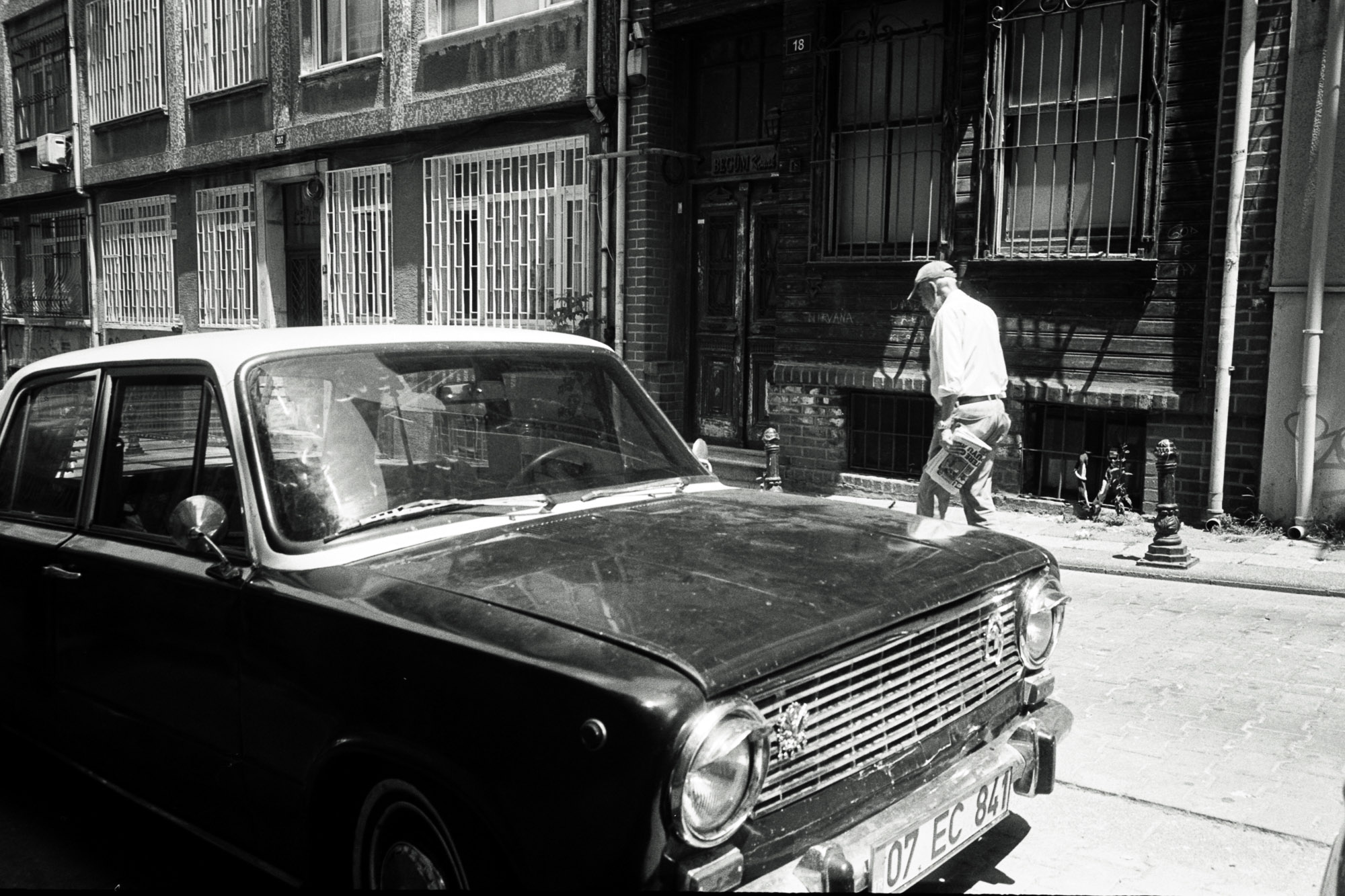 A black and white vintage car is parked on a narrow cobblestone street beside a brick building with barred windows, as a person walks by holding a newspaper in the background.