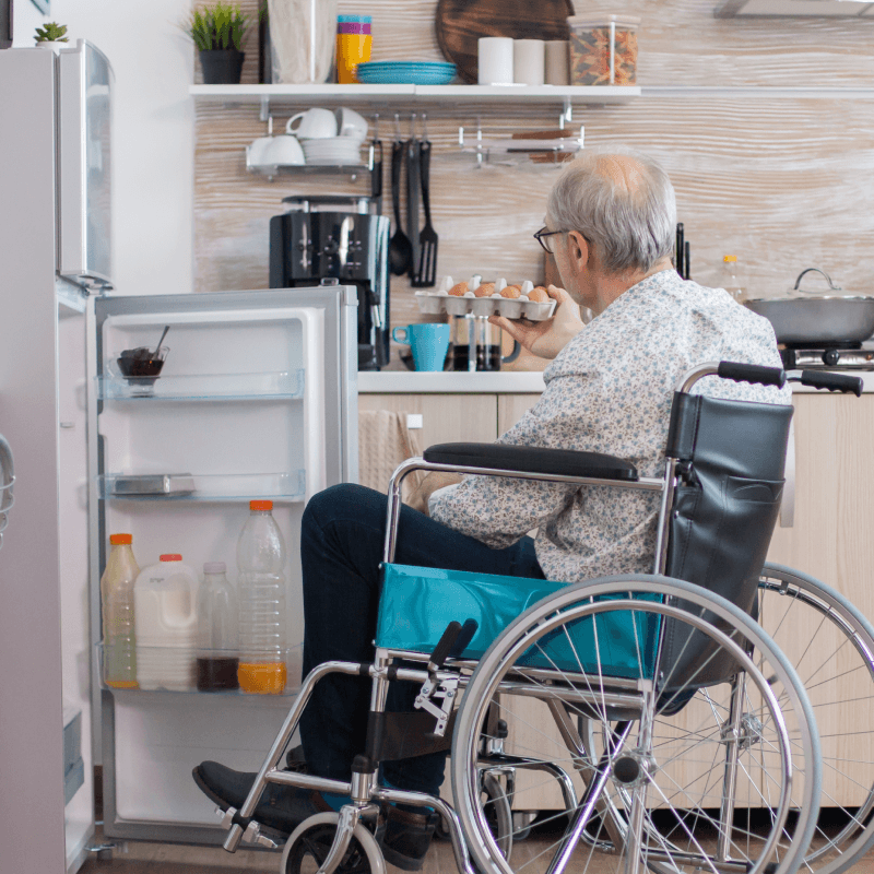 An older man in a wheelchair sits in a kitchen, holding an egg and facing an open refrigerator with bottles and containers on the door shelves.
