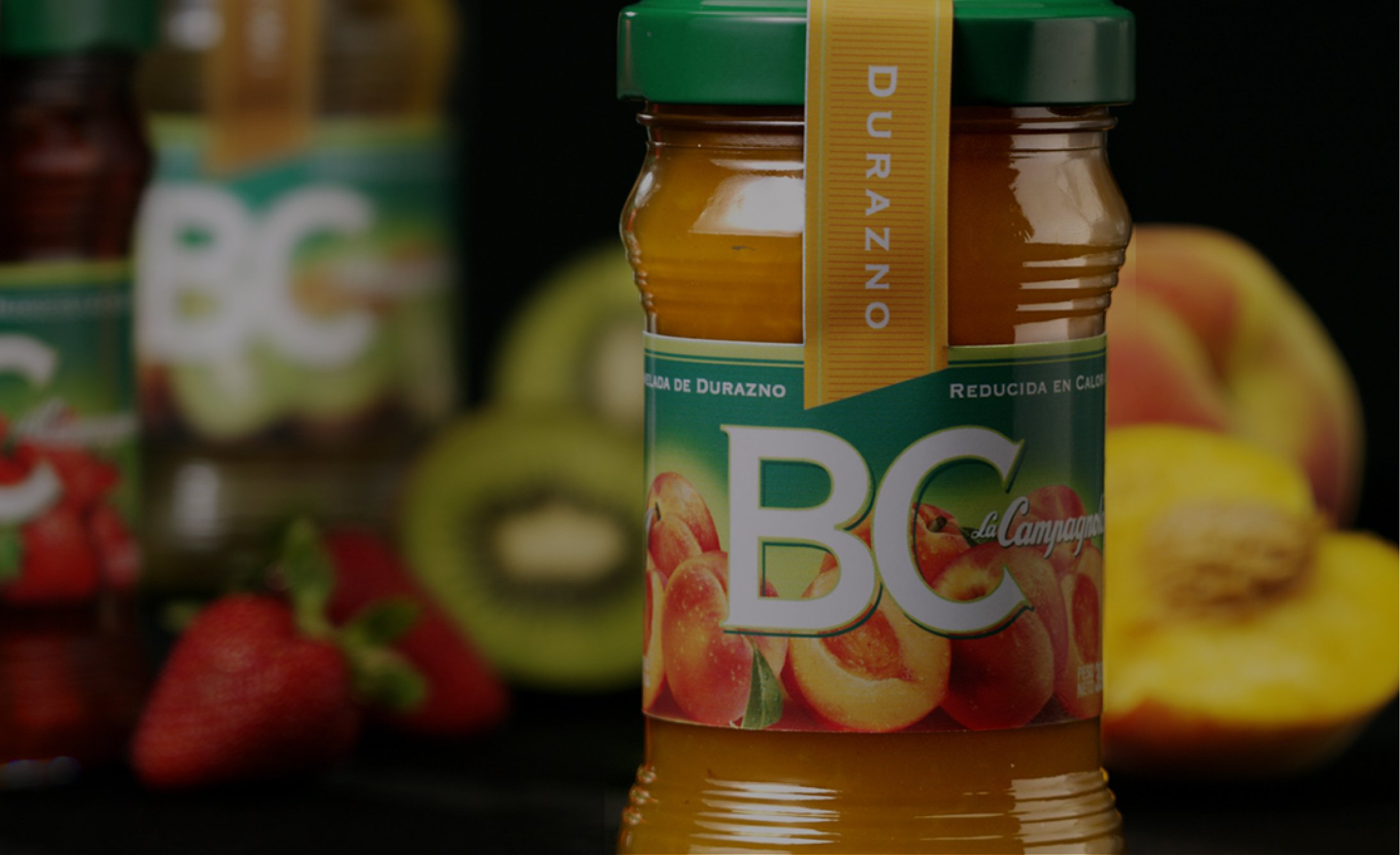 Jar of peach jam labeled "BC" in the foreground, with strawberries, kiwi, and a blurred peach in the background, conveying a fresh, fruity theme.