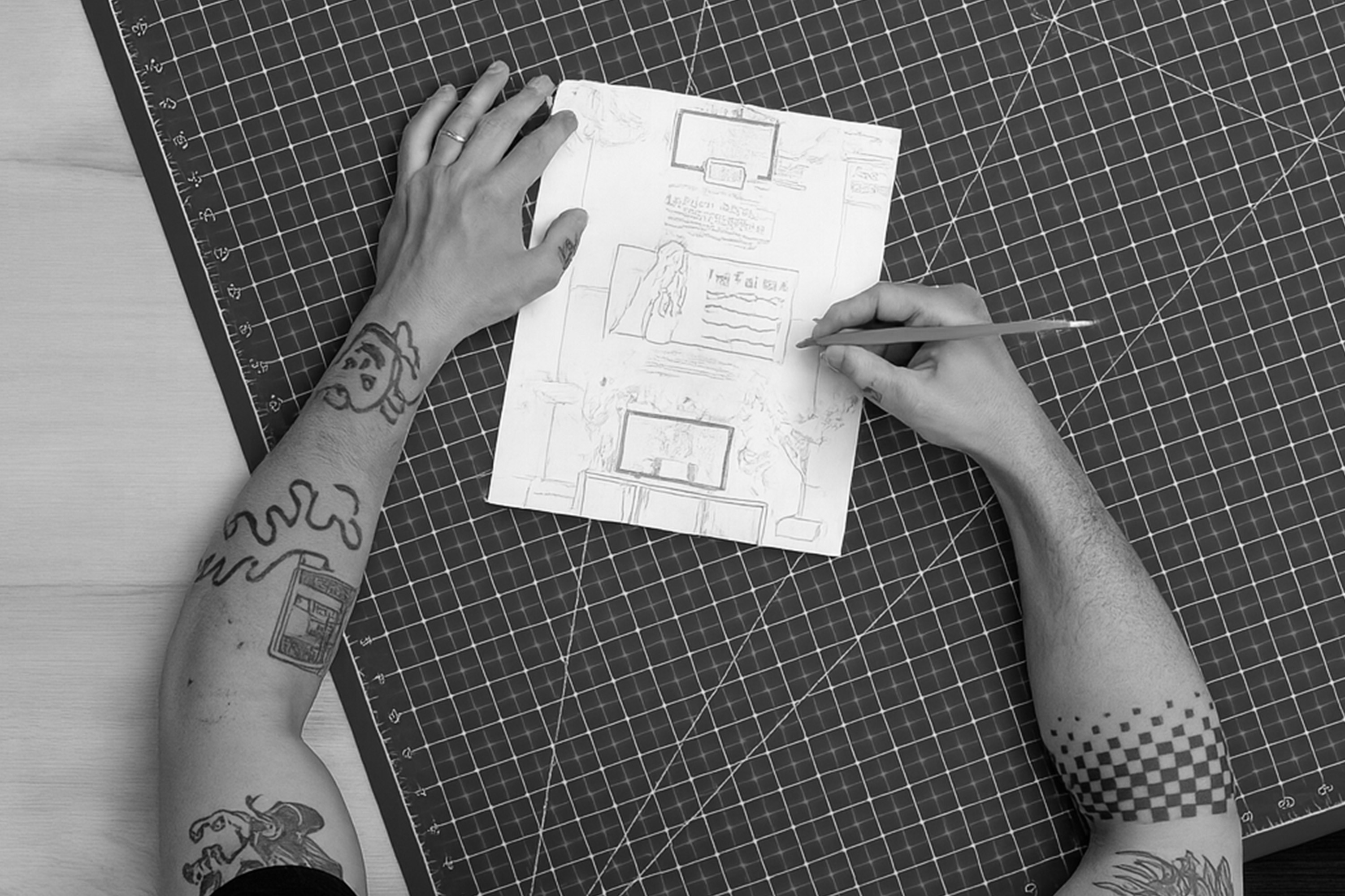 A bird's eye view of two tattooed arms holding and drawing on a piece of paper with a grid cutting mat underneath.