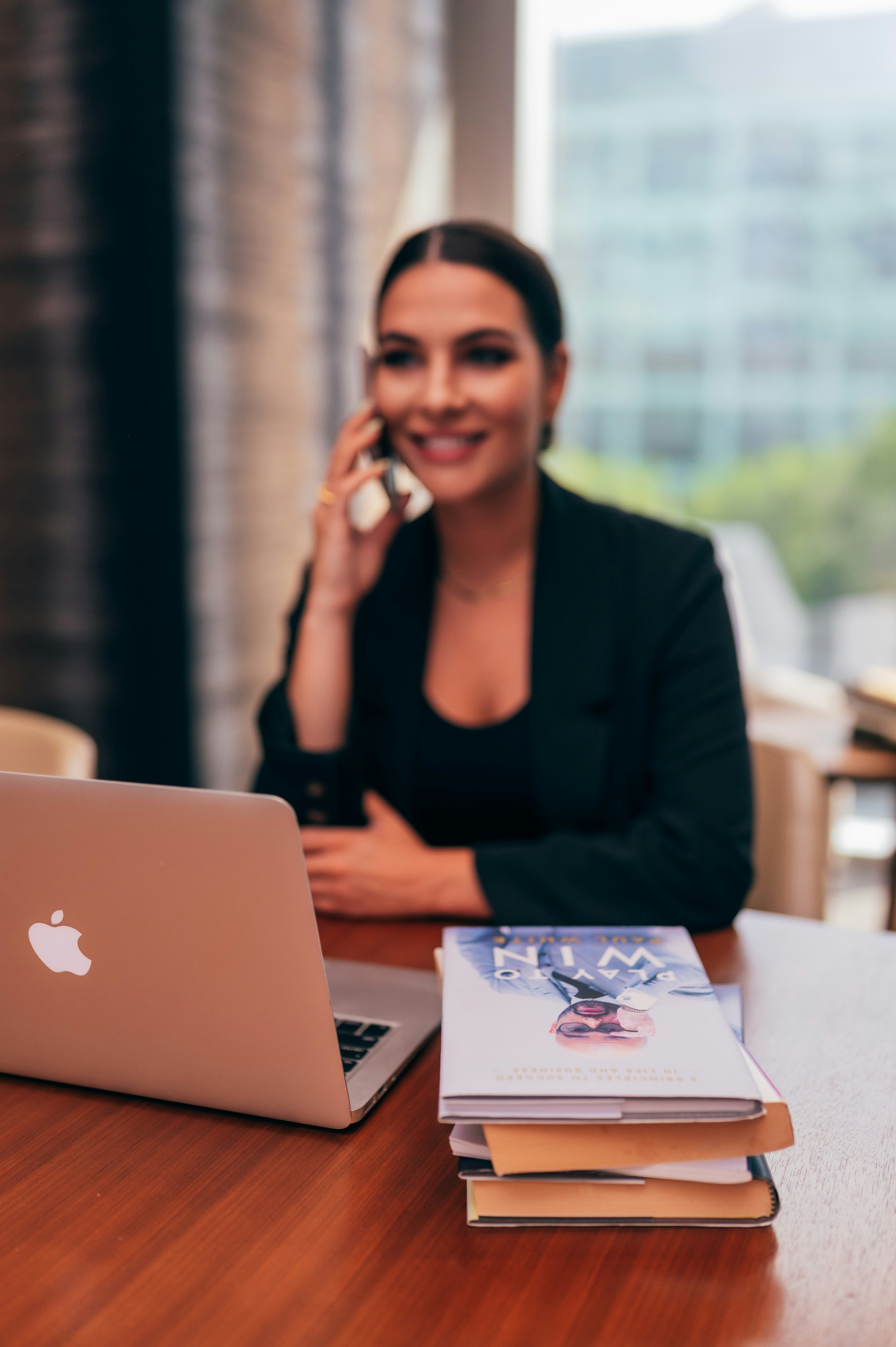 Mulher com cabelos presos e roupra sosical preta usando celular sentada de frente para mesa com um notebook