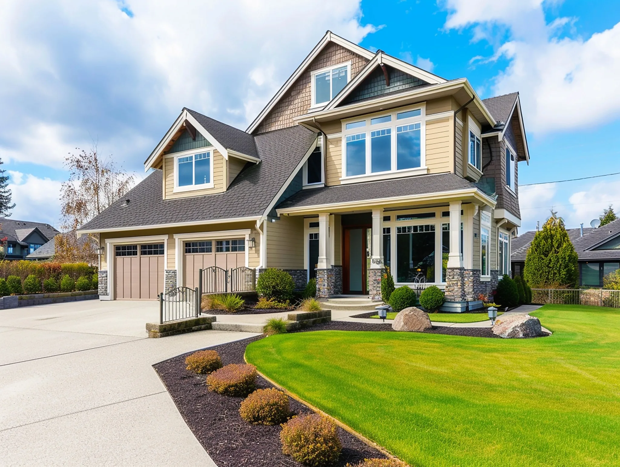 A modern two-story suburban home with cream siding and dark gray roof features a two-car garage, front porch with stone pillars, large windows, and well-manicured landscaping including green lawn and decorative shrubs under a partly cloudy blue sky.
