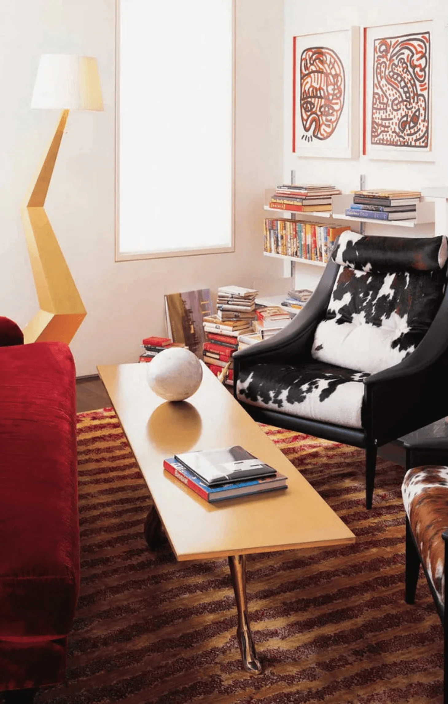 A red velvet sofa and cowhide armchair sit beside a gold coffee table and angular brass floor lamp, with two Keith Haring prints on the wall