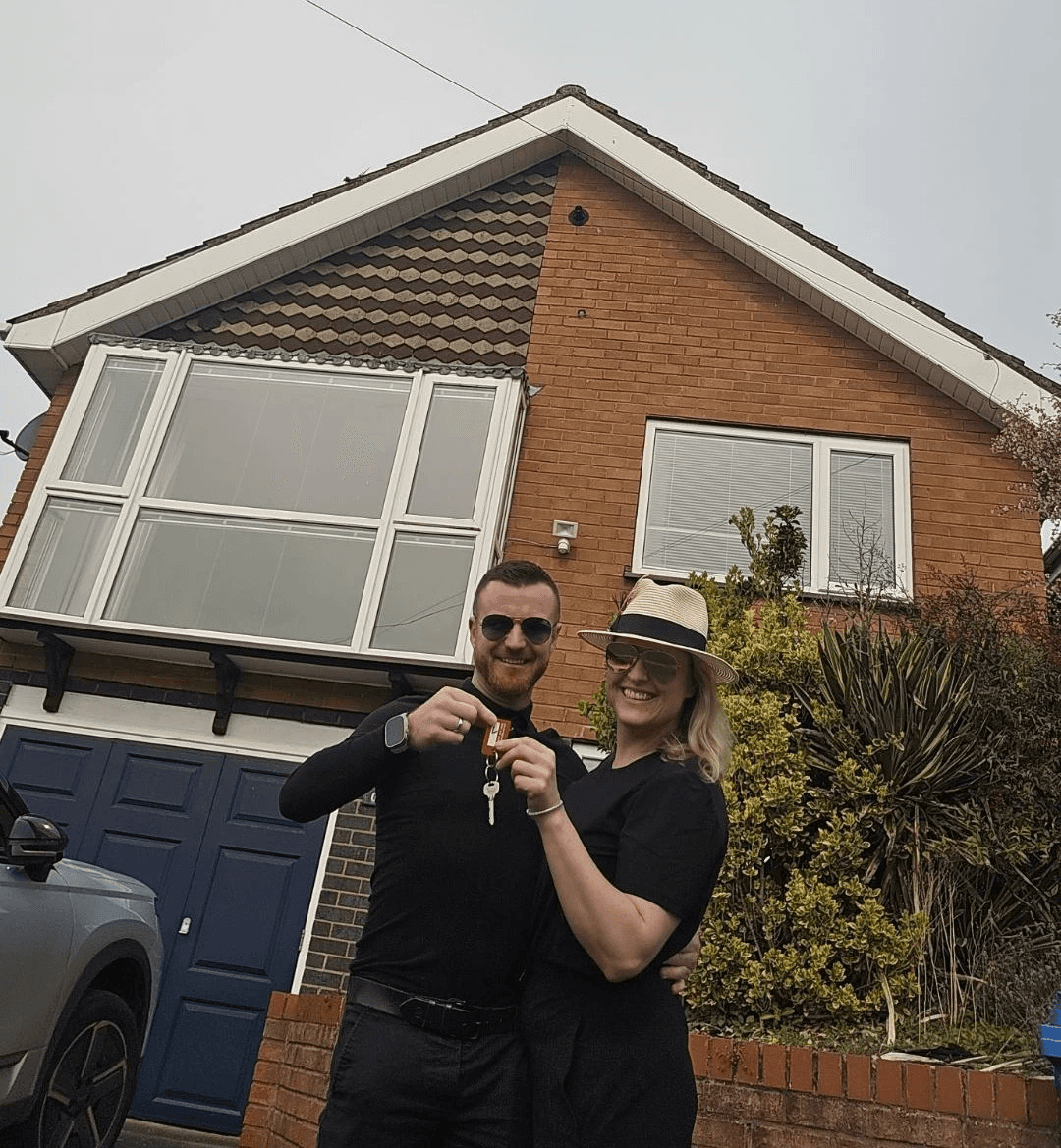 Man in grey jacket smiling and holding up house keys in front of a brick house entrance