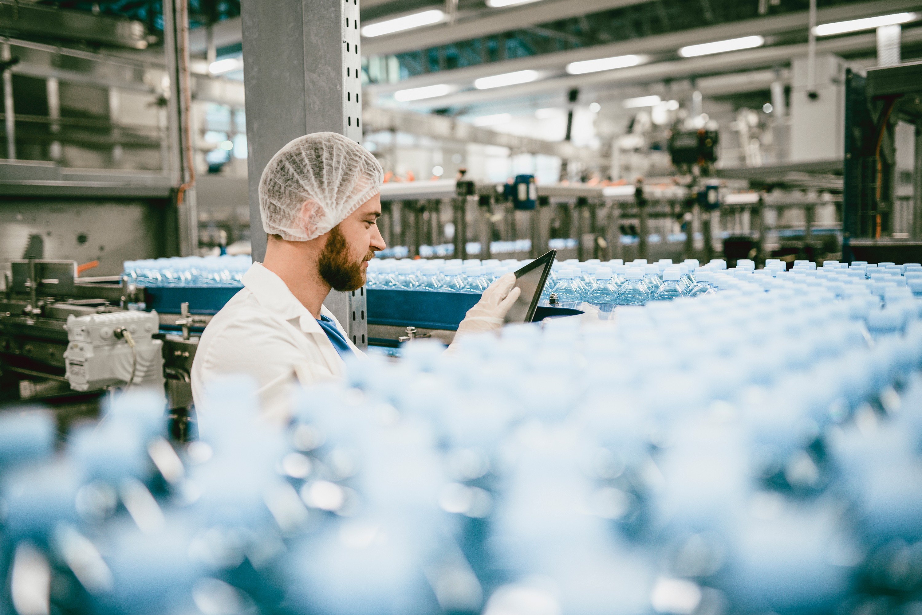Man in a hairnet, white coat and gloves checking a tablet computer on a bottling line filled with rows of blue plastic bottles