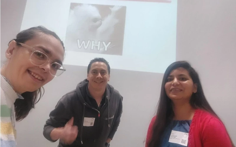Three people stand together at an indoor event space with a presentation slide displayed behind them reading “WHY.” Name badges and casual attire suggest a workshop or collaborative session.