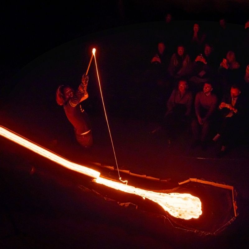 A Lava Show host uses a metal rod to interact with glowing molten lava flowing inside a dark showroom, while seated guests watch the live demonstration illuminated by the bright orange lava.