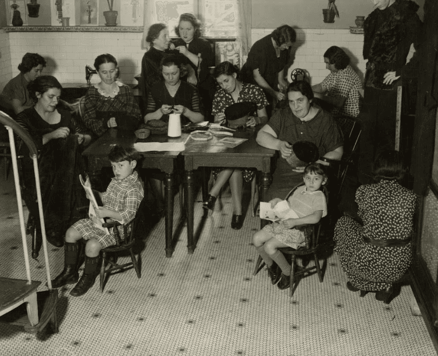 Women and children from the 1930s sit in a small kitchen working on crafts