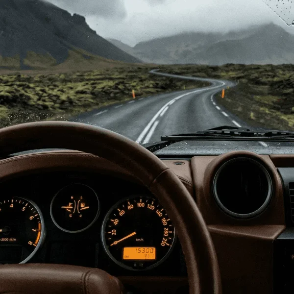 View from inside a car with a brown dashboard and gauges, looking through a rain-spotted windshield at a misty mountain landscape.