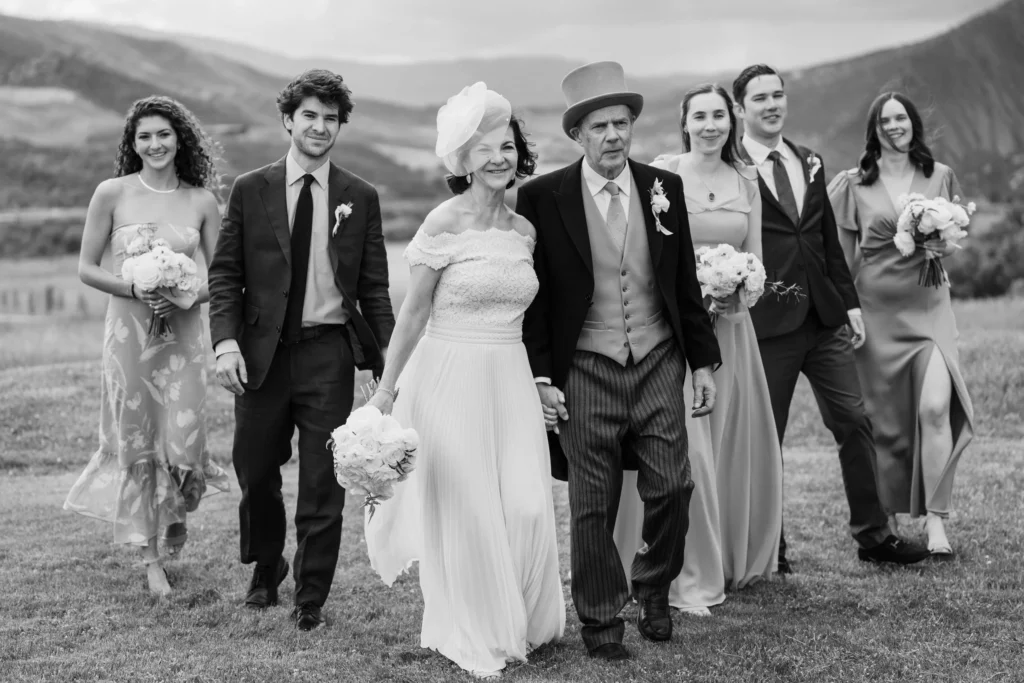 A black and white group photo of the wedding party walking across a field with mountains in the background at a Chaparral Ranch Wedding.