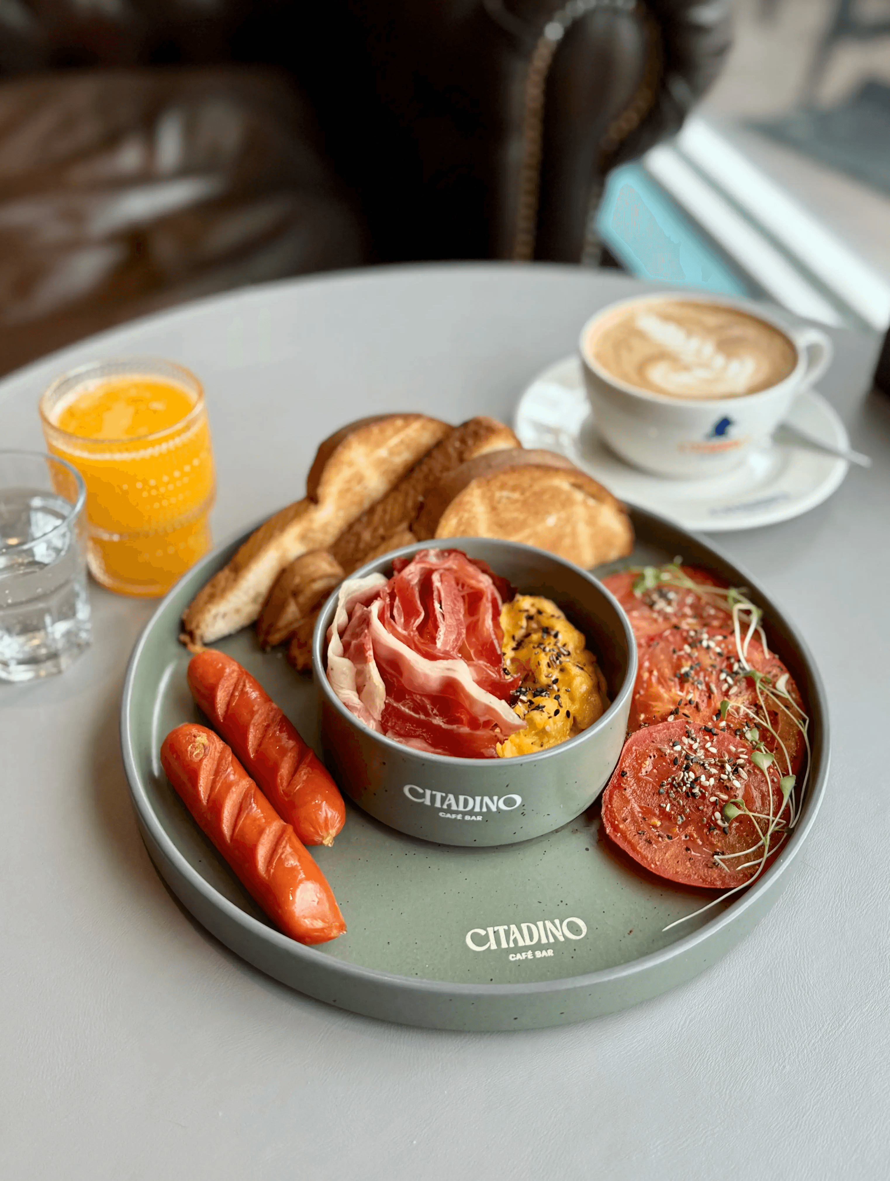 desayuno con salchichas, tostadas, jamón crudo, huevos revueltos con semillas, jugo de naranja, agua y café.  