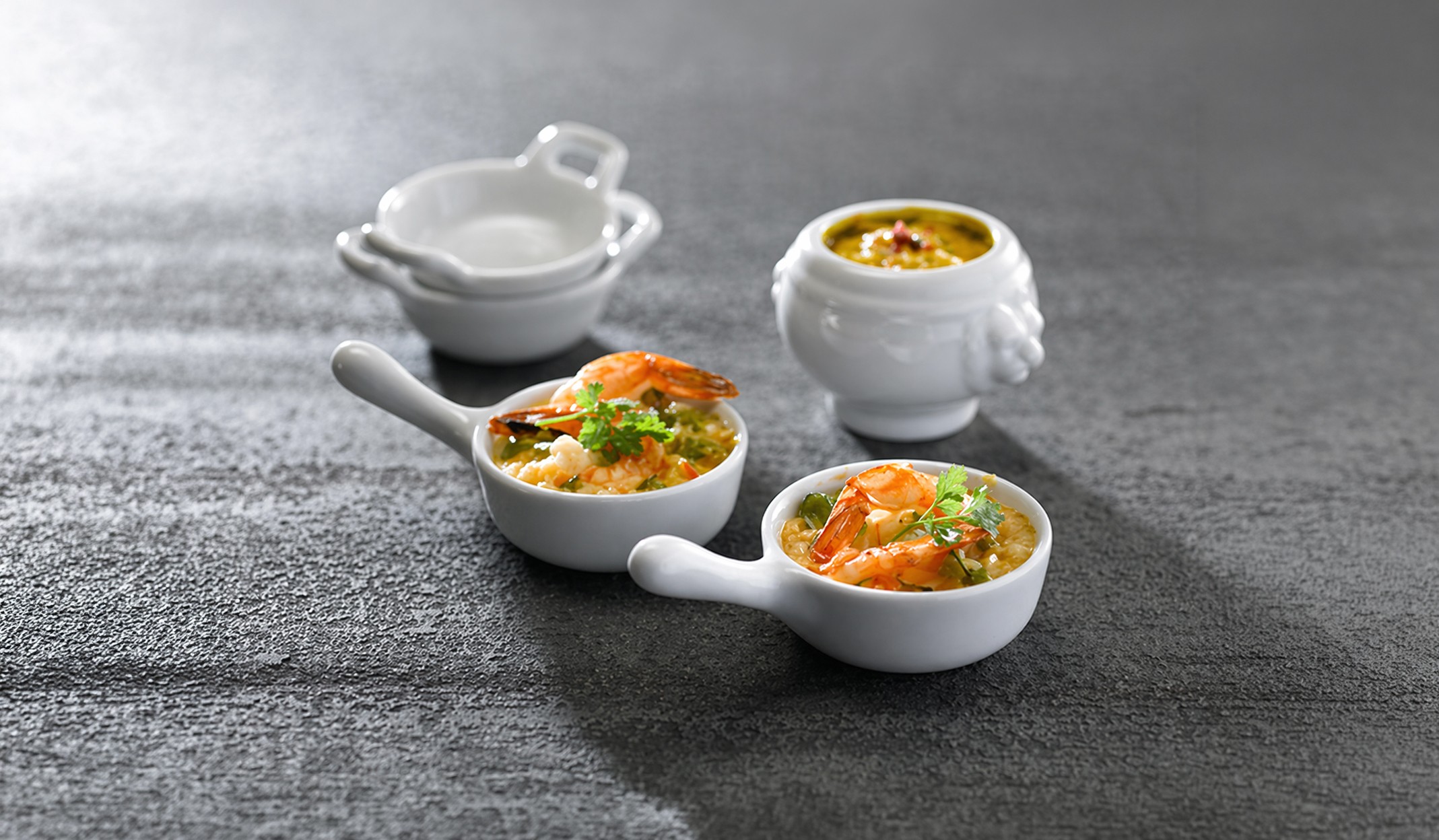 Assortiment de coupelles et mini-cocottes en céramique blanche présentées avec des gambas et une soupe, sur fond gris texturé