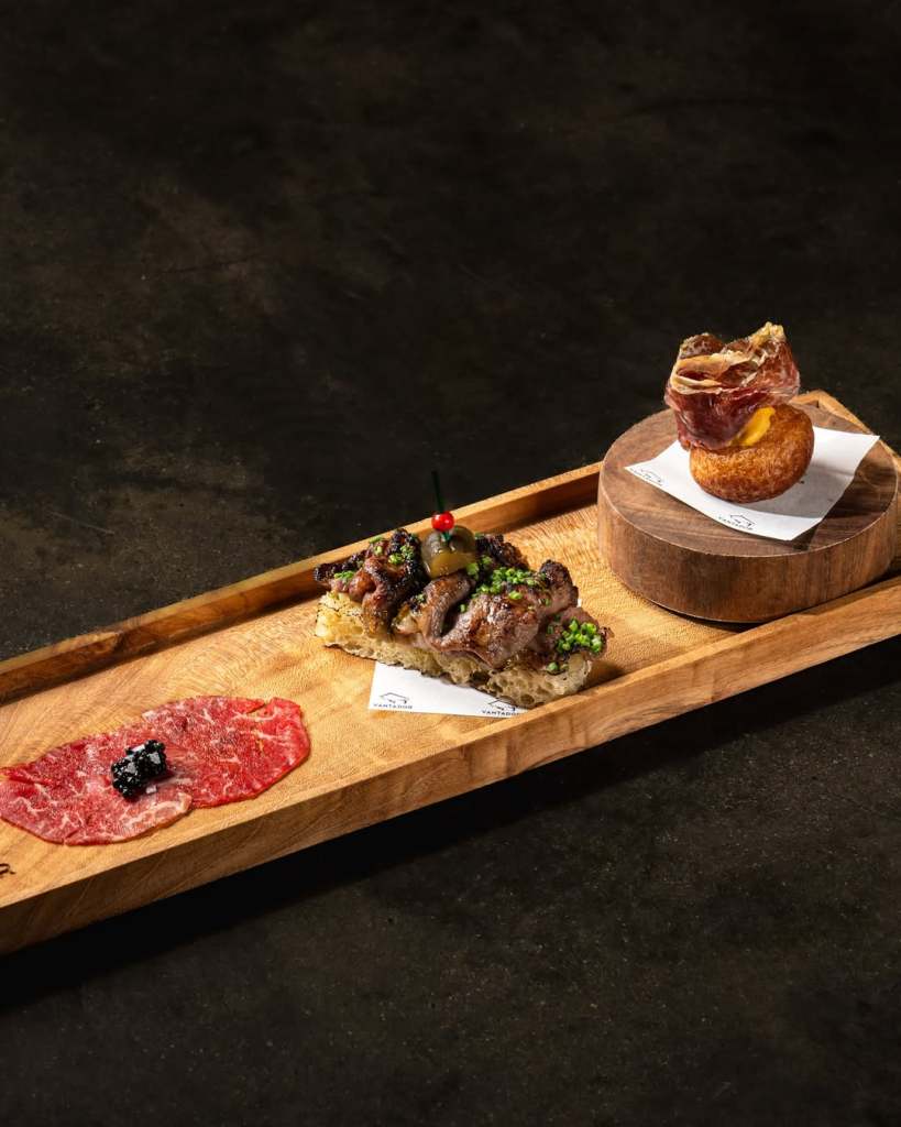 A wooden platter displaying a gourmet appetizer selection, featuring a slice of cured meat topped with caviar, a grilled meat dish garnished with herbs and a cherry tomato, and a mantao donut filled with cheese and topped with cured beef.