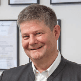 Hubert Zeltsperger steht im Anzug vor einer Wand mit Zertifikaten und lächelt